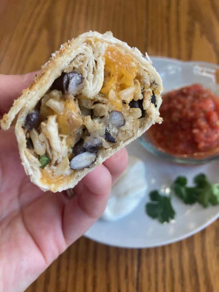 Hand holding a crispy chicken, rice and bean burrito in front of a plate with salsa and sour cream.